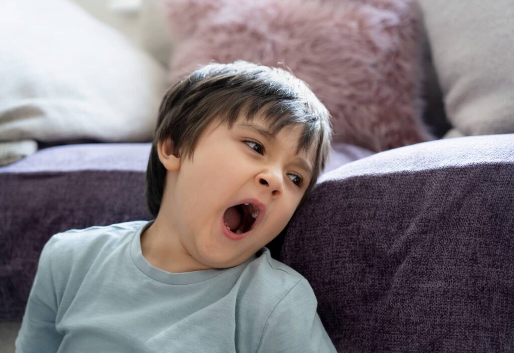 An image of a tired kid yawning sitting next to sofa.
