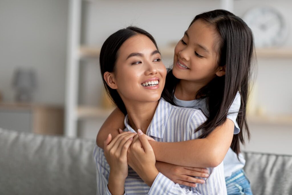 An image of a mom and daughter hugging and smiling, girl embracing mother from back, enjoying spending time together at home.