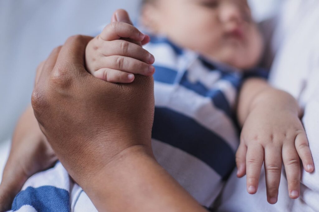 An image of a father's hand holding baby hand with blurry image of sleeping baby in a striped blue bodysuit.