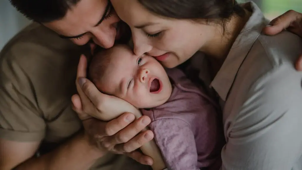 An image of young parents holding and kissing their newborn baby indoors at home.