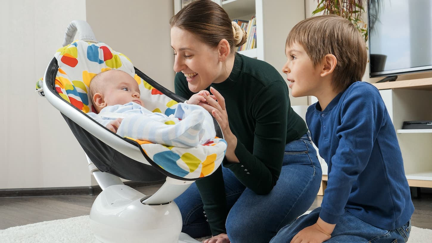 An image of a baby in a mamaroo with mom and sibling looking on.