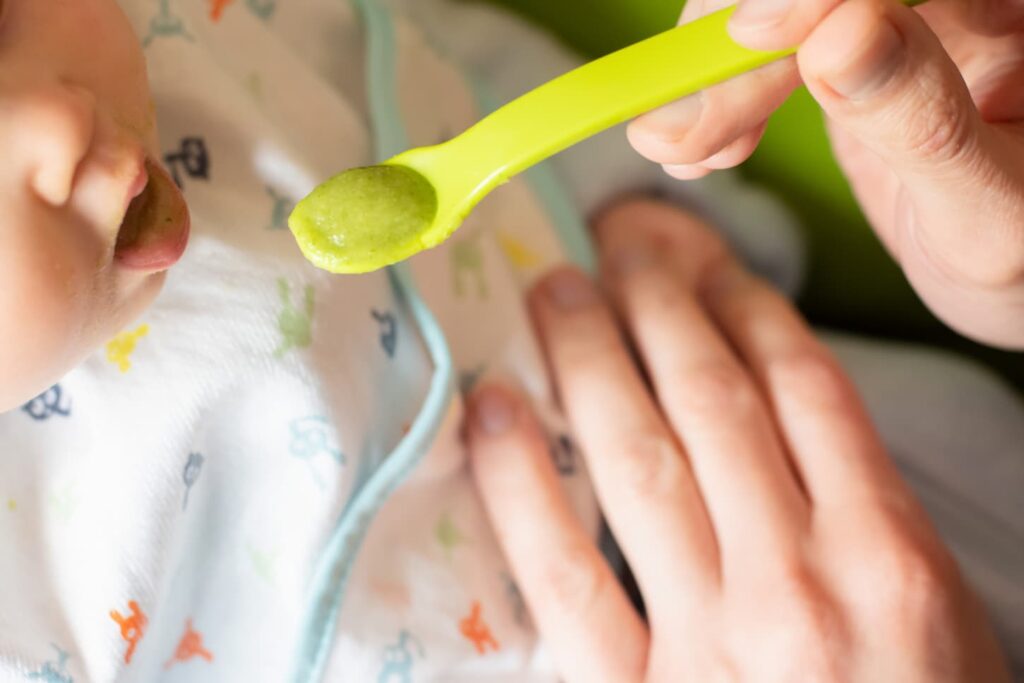 An image of an adult feeding pureed broccoli to a little baby.