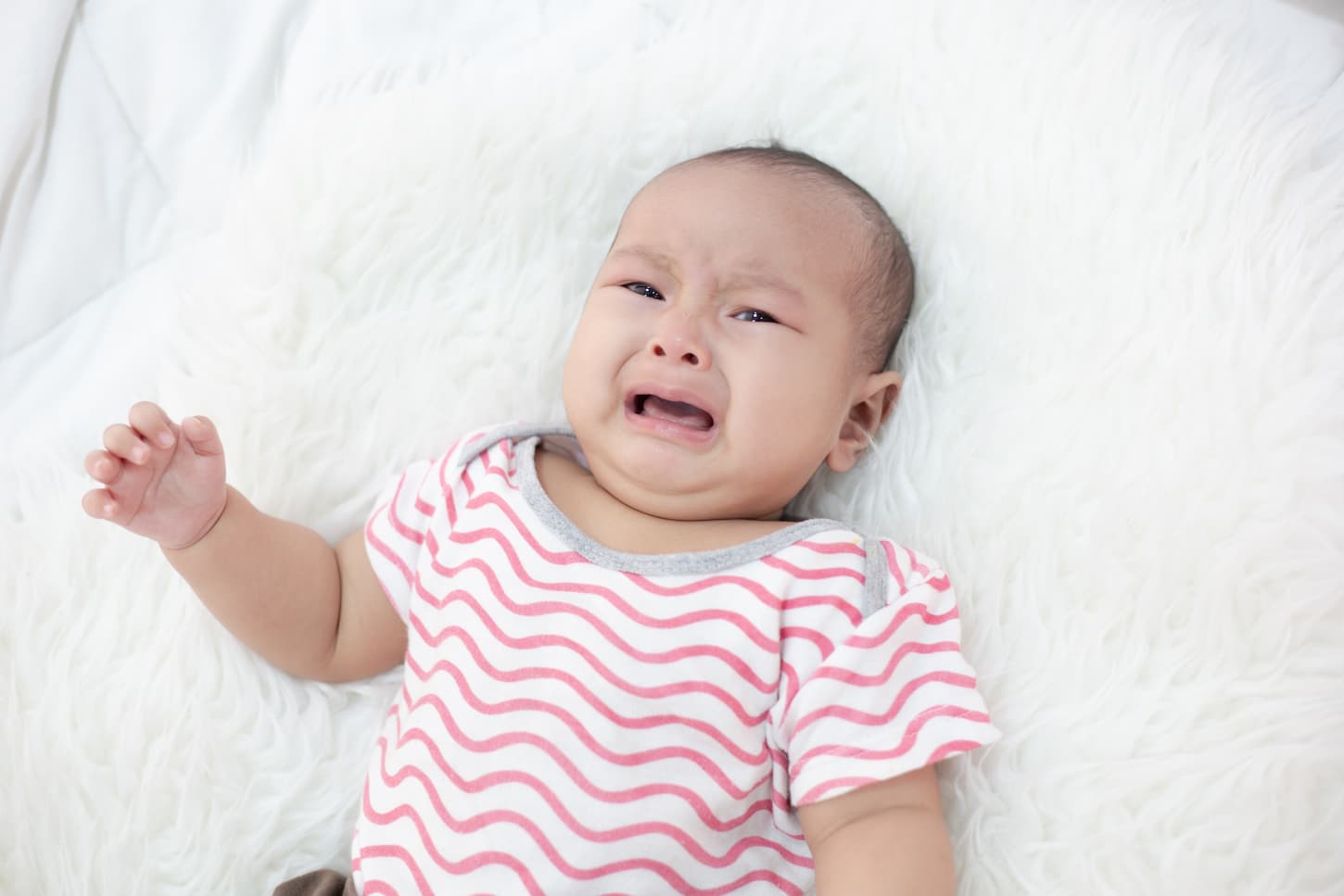 An image of a baby girl wearing a pink-striped short sleeve baby suit while crying in a crib.