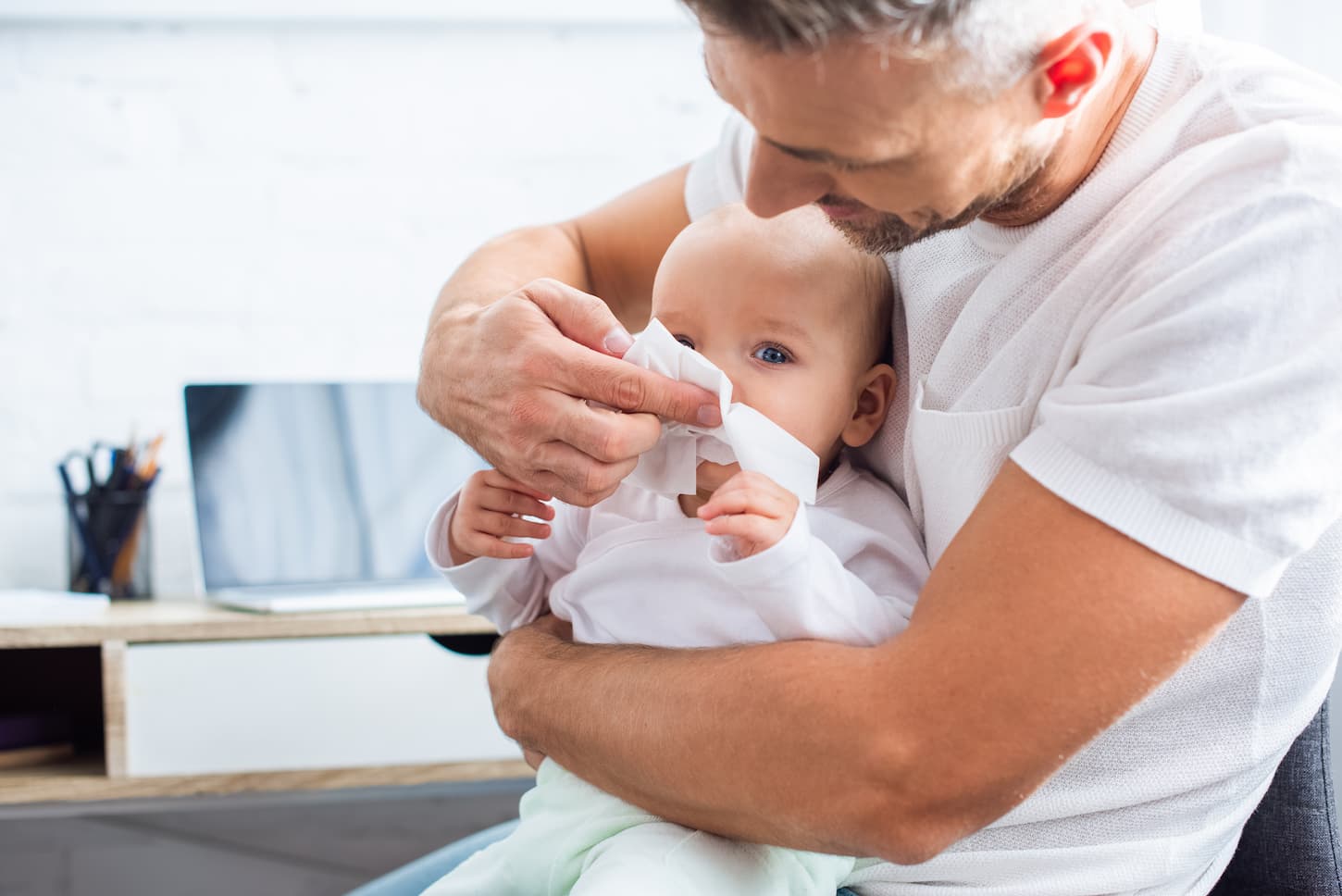 An image of a father wiping the runny nose of an adorable baby daughter with a napkin at home.
