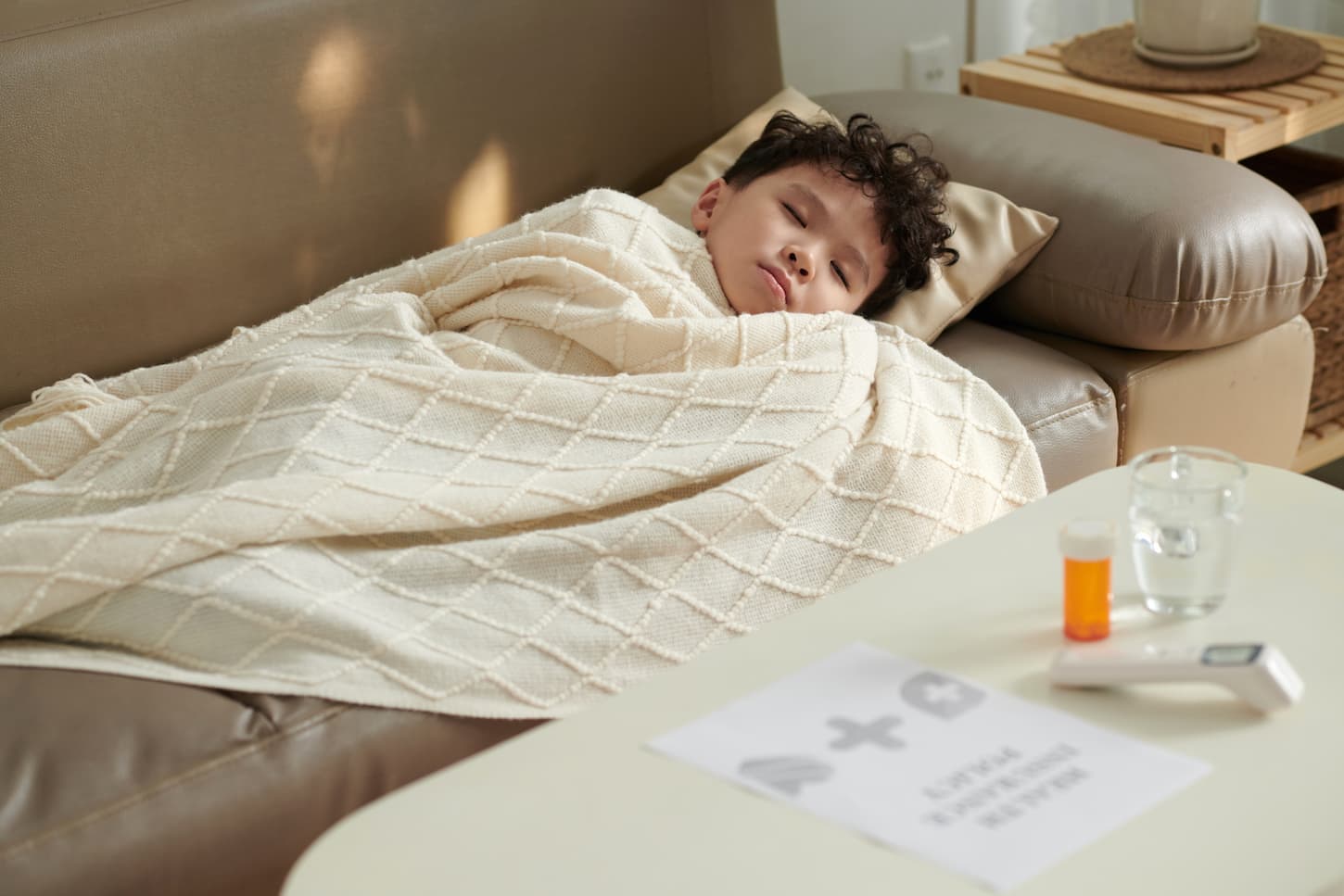 An image of a little sick boy tucked in a blanket sleeping with medicine, a glass of water, and a thermometer on a table near the sofa where the little boy sleeps.
