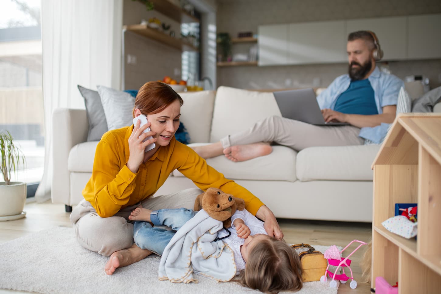 An image of a child laying on the floor while her mother is on the phone and her father in front of a laptop working at home.