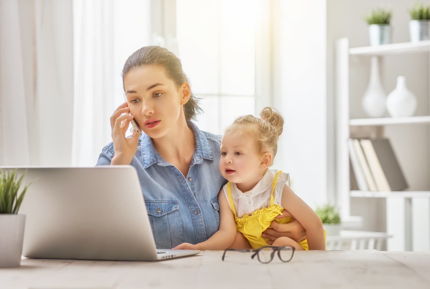 An image of a mother talking with someone over the phone while looking at her working laptop and holding her adorable daughter in another hand.