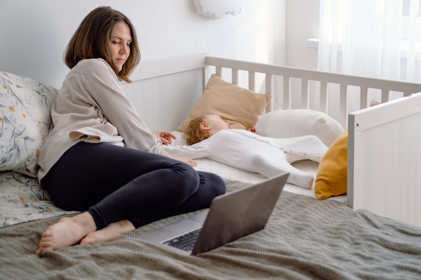 An image of a mother working on a computer or laptop while her baby is taking a daytime nap in a baby cot.