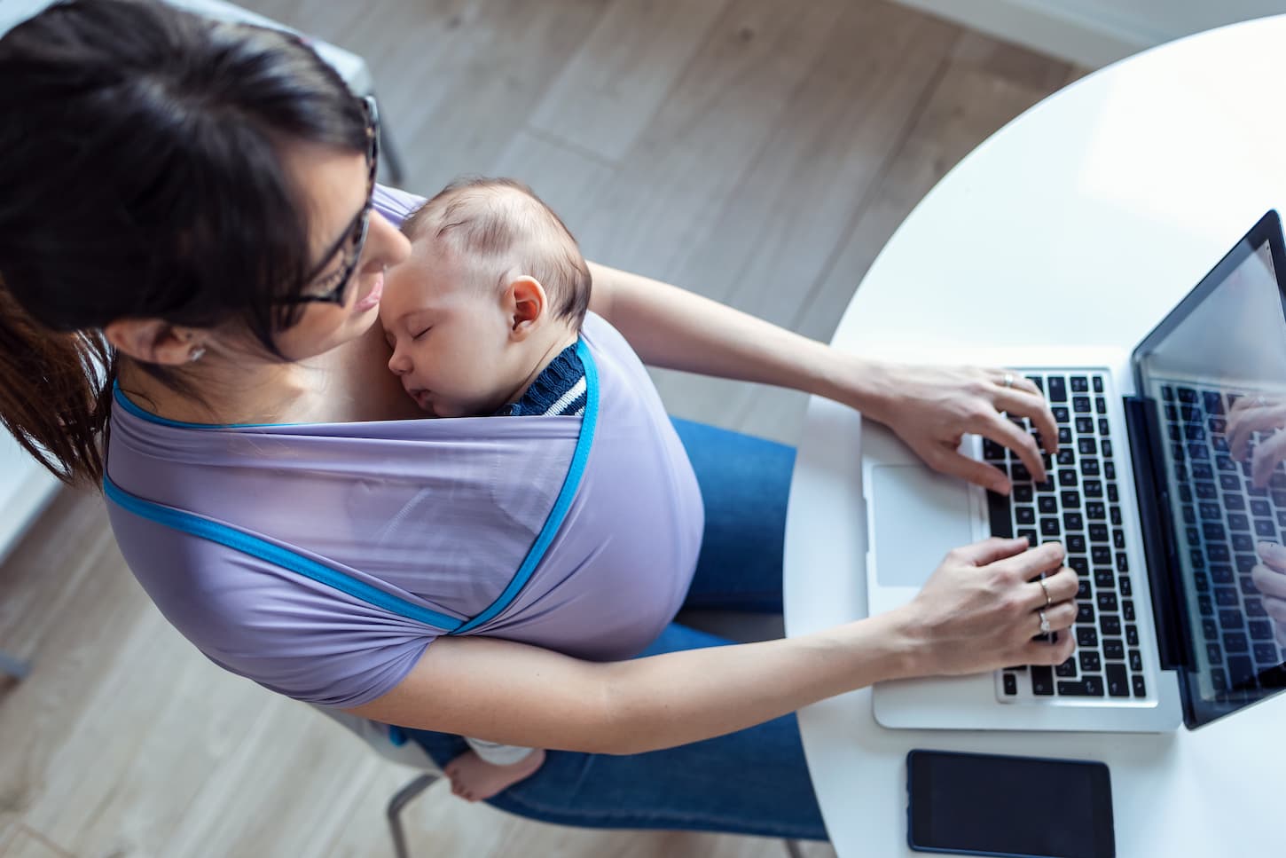 An image of a pretty young mother with her baby in a sling working with a laptop at home.