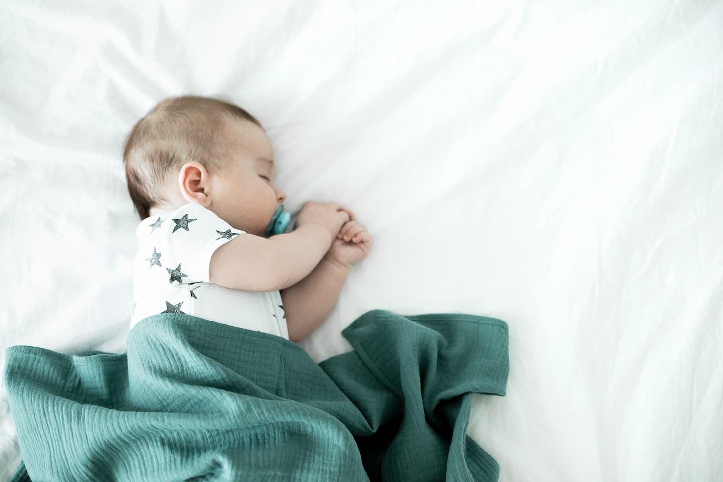 An image of a 3-month-old baby sucking a pacifier and sleeping alone on a bed covered with a green blanket.