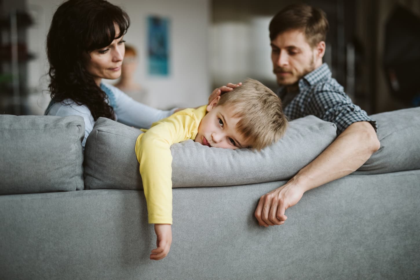 An image of a bored young kid lying on a couch next to his young worried parents.