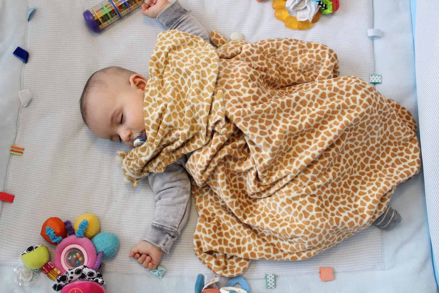An image of a Sleeping baby boy in the playpen with a blanket and pacifier in his mouth.