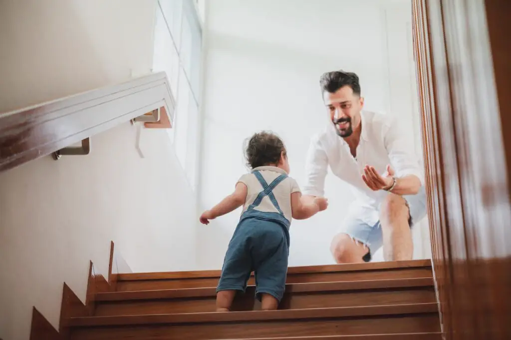 An image of a Handsome young father playing with little child daughter who is climbing stairs at home and man motivating her to come upstairs.
