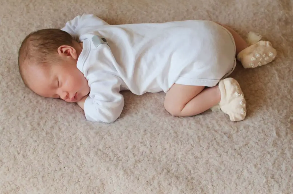An image of a newborn sleeping sweetly on his tummy in full-length white clothes in a mattress.