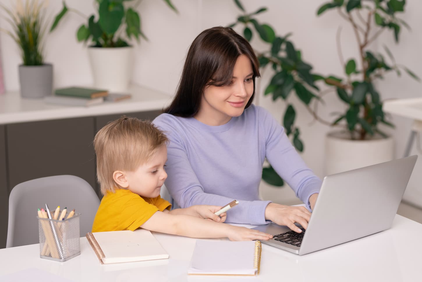 An image of a Cute son sitting at the table and touching a mother's laptop while assisting her with work, a young mom working at home.