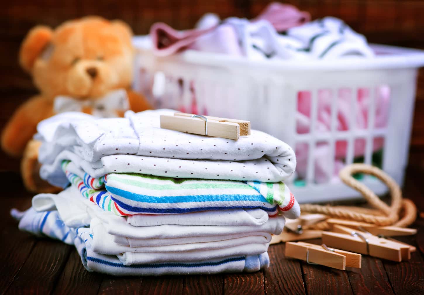 An image of baby clothes on the wooden table with a teddy bear and a basket on the blurred background.