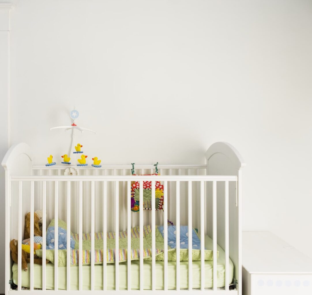 An image of a white baby crib consists of comfortable blankets, toys, and baby mobile for the crib.