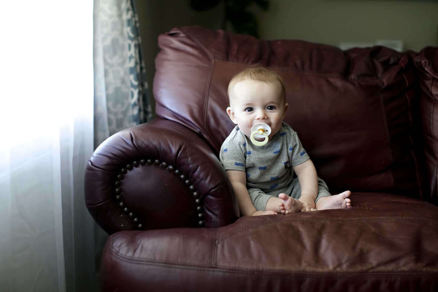 An image of a baby sitting on a brown leather sofa with a pacifier while looking at a camera.