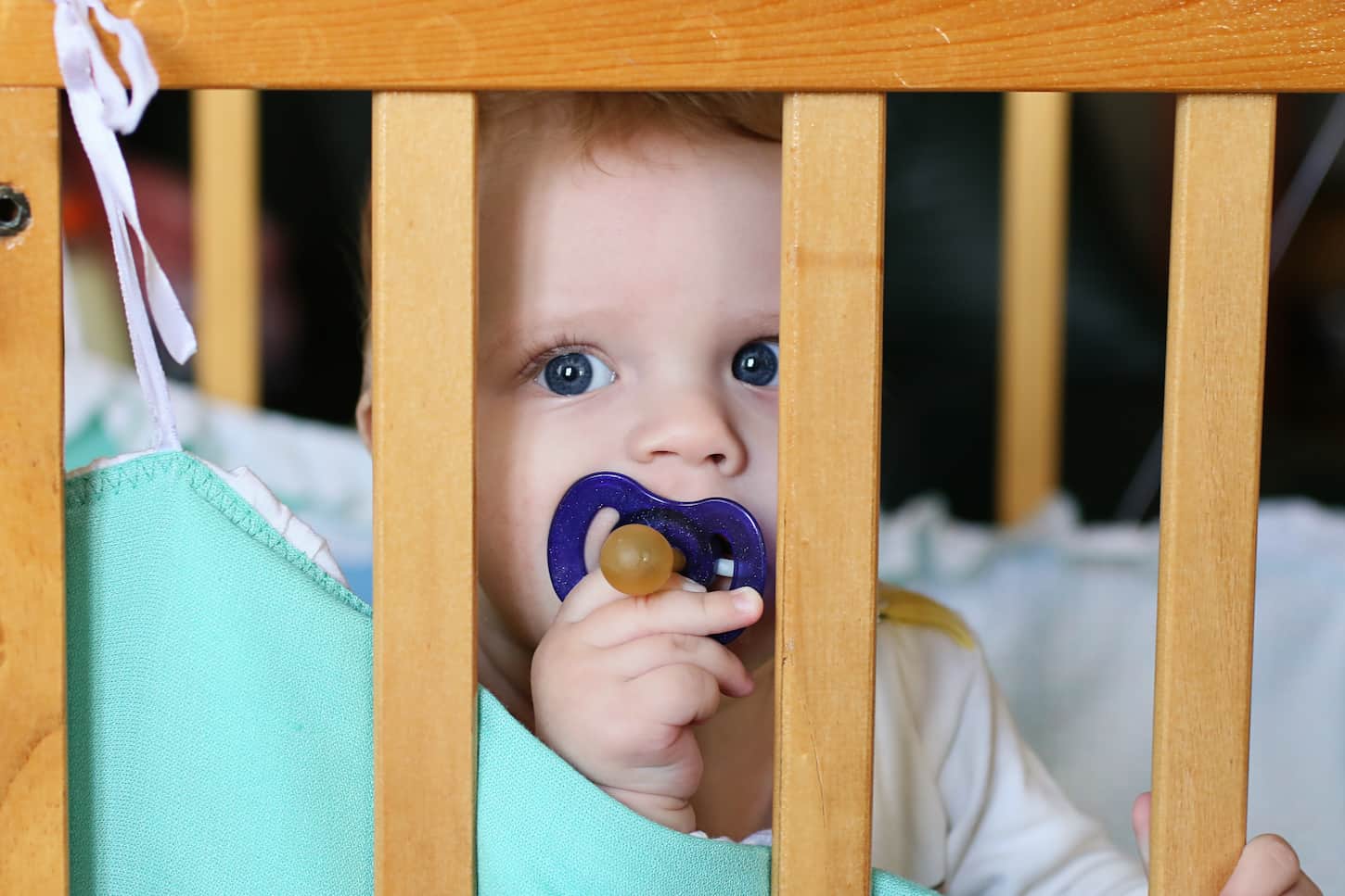 An image of a cute infant holding his pacifier and sitting in his bed.