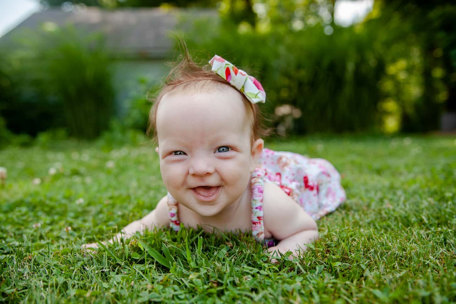 An image of a Four-month-old infant girl wearing a pink floral print romper and hairbow doing tummy time outside in the yard on a warm day.