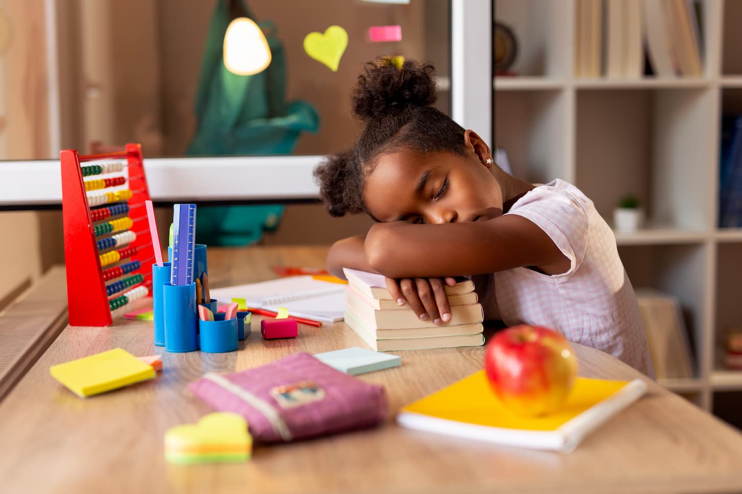 An image of a beautiful little girl tired of doing her homework, sleeping at her desk, using a pile of books as a pillow.