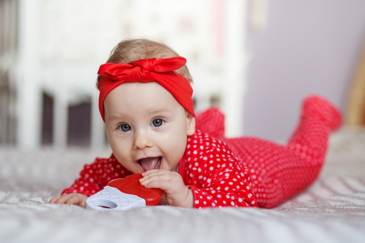 An image of a Little 6 months old baby in a red sleepsuit and headband lying on the bed on her stomach.