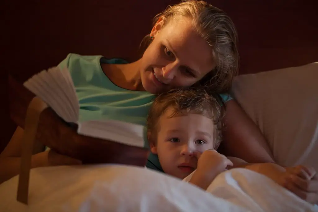 An image of a mother reading a bedtime story to her little son cuddling down alongside him on the bed as he peers over the counterpane at the camera.