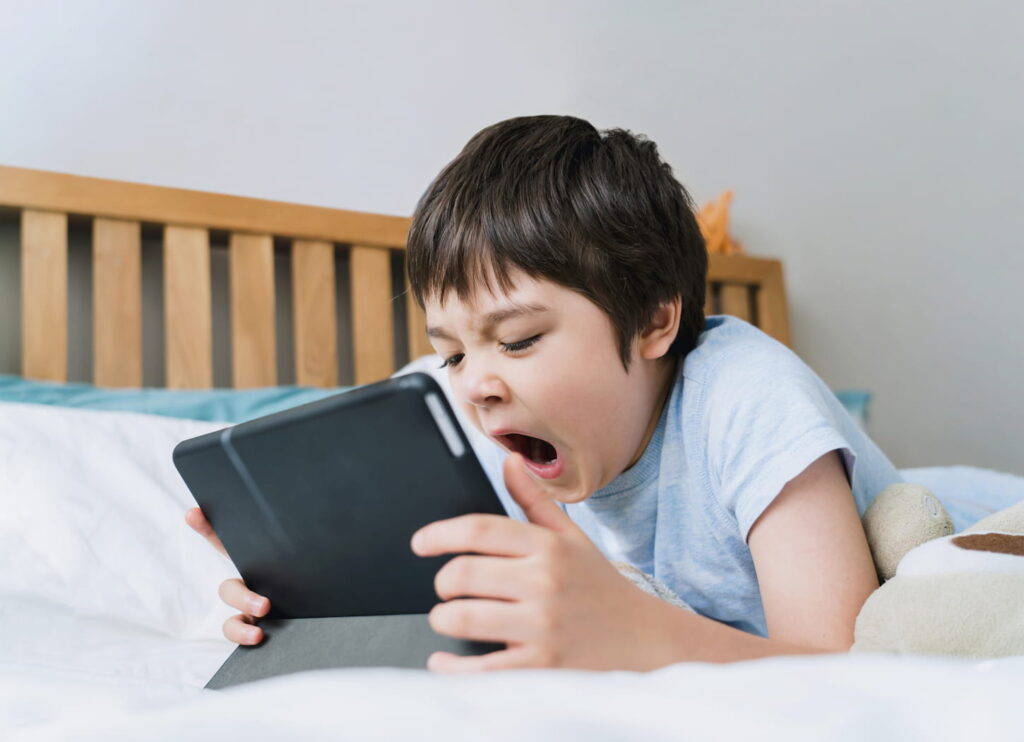 An image of a tired sleepy boy yawning and lying in bed in the morning while watching cartoons or playing games on a tablet in the bedroom.