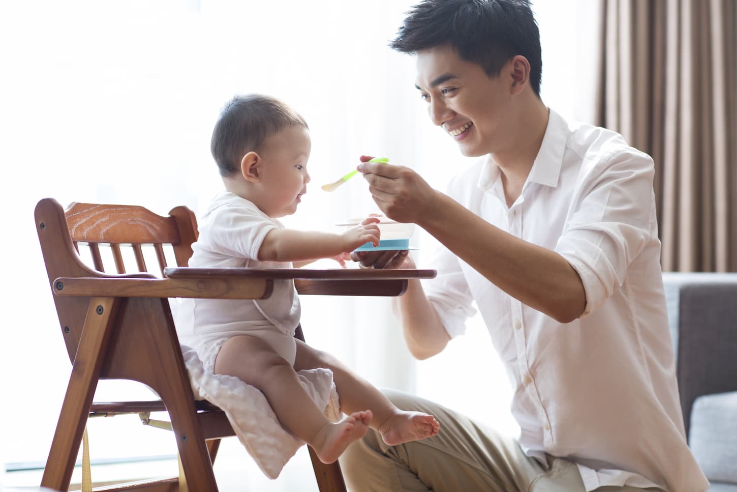 An image of a Young father feeding a baby in a high chair.