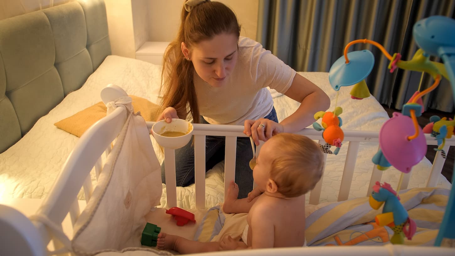 An image of a young mother giving soup with a spoon to her baby son sitting in the crib at night.