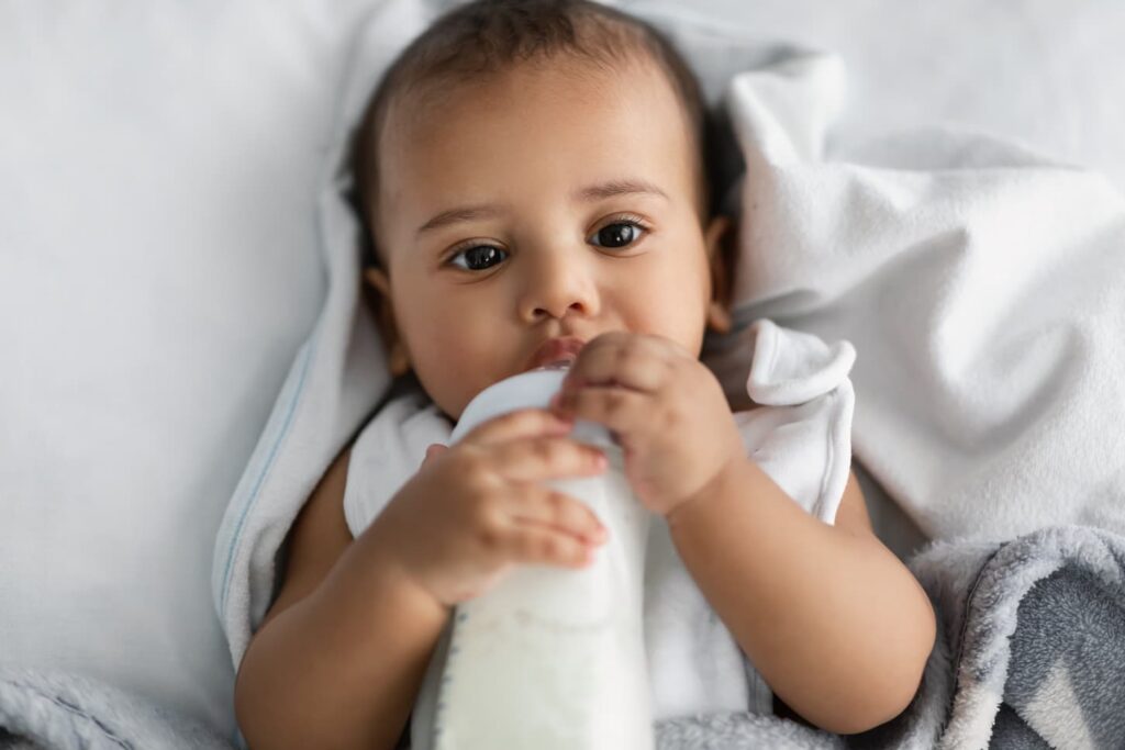 An image of an adorable small African American baby drinking milk from bottle, lying on the white blanket in crib.
