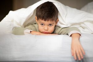 An image of a Tired sad caucasian little child lies on bed, under blanket, looks at camera in bedroom, having Problems with sleep.